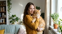 Young Caucasian woman in yellow sweater holding orange tabby cat in cozy living room with plants and natural light creating warm domestic atmosphere.