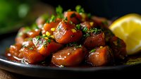 Fresh salmon poke cubes glazed with soy sauce and sesame, garnished with green onions and cilantro on dark plate with lemon wedge. Asian cuisine appetizer.