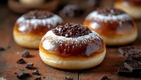 Fresh baked chocolate donuts dusted with powdered sugar on rustic wooden surface with chocolate chunks scattered around, close up view of sweet pastries.