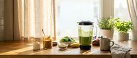 Fresh green smoothie in blender on sunny kitchen counter with healthy ingredients and plants creating morning wellness lifestyle scene.