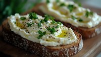 Fresh ricotta cheese spread on artisan bread slices with herbs and olive oil drizzle. Healthy breakfast or appetizer concept for restaurant menu design.