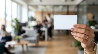 Mature male professional holding blank white business card in modern office environment with colleagues working in background, suitable for corporate branding and business communication concepts.
