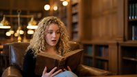 Young blonde woman with curly hair reading book in cozy library with warm lighting and wooden shelves creating studious atmosphere for education content.
