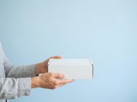 Male hands holding white cardboard box against blue background for product delivery, shipping, unboxing, e-commerce, and retail packaging concepts.