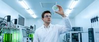 Young Caucasian male scientist in white lab coat examining test tube with green liquid in modern laboratory with advanced equipment and bright lighting.
