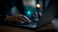 Glowing atomic symbol hologram projection over laptop keyboard while person types in dark room, representing quantum computing and future technology.