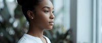 Young African American businesswoman in white shirt looking thoughtfully out office window during daytime, professional corporate environment.