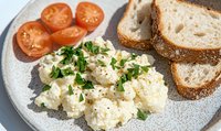 Creamy scrambled eggs garnished with fresh parsley served on gray plate with sliced cherry tomatoes and whole grain bread. Classic breakfast setting.