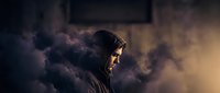 Mysterious hooded figure emerges from dramatic smoke and fog with cinematic lighting creating atmospheric mood for thriller and suspense projects.