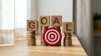 Wooden blocks spelling GOAL with red target symbol and coin stacks on table representing financial planning and achievement motivation for business success.