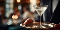 Elegant martini cocktail served on silver tray by waiter in upscale bar with atmospheric bokeh lighting in background.