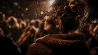 Young woman with messy bun hairstyle in brown teddy coat watching concert from crowd, with bokeh lights and falling snow creating atmospheric nighttime ambiance.