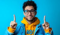 Young Hispanic man with curly hair and glasses wearing yellow hoodie and blue jacket points fingers up while smiling against turquoise background. Positive expression.