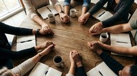 Business team joining hands together over wooden table during meeting with coffee cups and documents showing teamwork and collaboration concept.