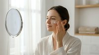 Young woman with East Asian ethnicity applying skincare cream to face while looking in mirror, demonstrating beauty routine and cosmetic product usage for wellness and personal care.