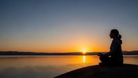 Woman silhouette meditating in lotus pose at serene lake during golden sunset with calm water reflection and peaceful mountain landscape horizon.