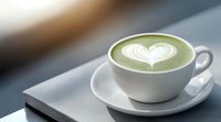 Green matcha latte with white heart-shaped foam art served in white ceramic cup and saucer on gray surface, soft natural lighting creates cozy atmosphere.