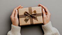 Female hands in cozy white sweater holding kraft paper gift box with elegant brown ribbon bow against neutral background, festive holiday present packaging.