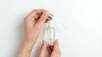 Hand holding clear glass dropper bottle with silver cap against white background for skincare and cosmetic product mockup.
