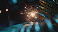 Bright sparkler emitting golden sparks against dark background with palm leaves silhouette, creating magical holiday atmosphere. Festive celebration moment.