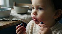 Toddler with dark hair and light skin eating fresh blueberries at wooden table, natural daylight kitchen setting, healthy snacking and childhood nutrition concept.