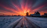 Dramatic winter sunset over snowy rural road with colorful sky rays illuminating countryside houses and bare trees in peaceful village landscape.