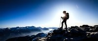 Male hiker with backpack standing on rocky mountain peak at sunrise, overlooking vast alpine valley landscape below with snow-capped peaks in distance.