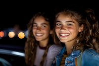 Two young Caucasian women with radiant smiles enjoying night city lights, wearing casual outfits. Natural authentic lifestyle portrait with bokeh background.