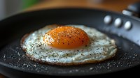 Macro shot of sunny side up egg with bright orange yolk and crispy edges, seasoned with black pepper in cast iron pan, morning breakfast cooking.