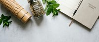 Natural herbal tea ingredients with dried herbs in glass jar, fresh mint leaves, wooden rolling pin and notebook on marble surface for wellness lifestyle.