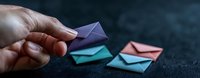 Hand holding small purple paper envelope with turquoise and coral origami envelopes on dark textured background, minimalist composition for correspondence concept.