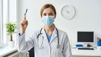 Female doctor in medical mask and white coat holding pen in modern clinic office with stethoscope around neck during healthcare consultation.