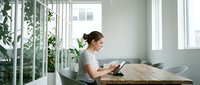 Young professional woman working on laptop at wooden desk in bright modern office with large windows and green plants creating productive workspace atmosphere.