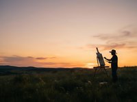 Male artist painting landscape during golden hour sunset in rural field with easel and canvas for creative work and outdoor art projects.