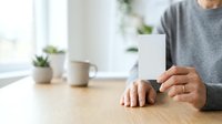 Man in gray shirt holding blank white card at wooden desk with coffee cup and plants for business messaging.