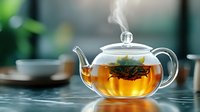 Glass teapot with brewing loose leaf tea and rising steam on marble table, warm amber liquid with floating tea leaves creates cozy atmosphere in soft focus.