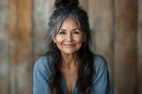 Mature Hispanic woman with long gray hair smiling warmly in casual blue shirt, representing aging gracefully and positive lifestyle for wellness content.