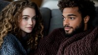 Young interracial couple relaxing on couch at home, wearing cozy knit sweaters. Woman with curly hair and man with beard sharing intimate moment.