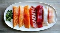 Fresh raw sashimi slices including salmon, tuna and white fish arranged on white oval plate with chopped herbs, showcasing gradient of colors from pink to red.
