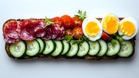 Open sandwich on dark bread with sliced salami, fresh cucumber, cherry tomatoes and boiled egg arranged in colorful rows, garnished with herbs.