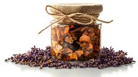 Glass jar with dried orange flowers and petals preserved in oil, decorated with burlap top and twine, surrounded by fresh purple lavender blooms on white surface.