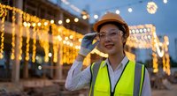 Young Asian female construction engineer in safety gear talking on phone at illuminated evening worksite with string lights and urban development background.