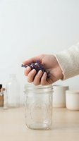 Hand holding dried lavender flowers over glass jar on wooden surface with skincare bottles and cosmetic containers in soft-focused background for wellness and natural beauty product marketing.