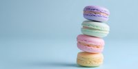 Stack of colorful French macarons in pastel purple, mint green, pink, and yellow against light blue background for food photography or bakery marketing.