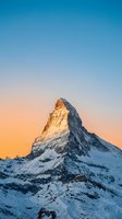Snow-covered alpine peak glowing with golden sunrise light against clear blue sky. Majestic mountain landscape for travel, nature, and winter adventure projects.