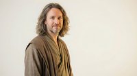 Middle-aged Caucasian man with shoulder-length wavy hair and beard wearing brown robe, smiling warmly against neutral background for wellness concepts.