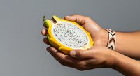 Young woman with medium skin tone holding halved dragon fruit with white flesh and black seeds against neutral background, suitable for food, nutrition, and healthy lifestyle content.