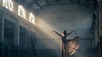 Young woman dancer performing graceful ballet movement in abandoned industrial warehouse with dramatic sunlight streaming through arched windows creating atmospheric mood.
