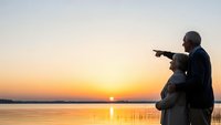 Senior couple silhouette pointing at sunset over calm lake water during golden hour romantic evening together creating peaceful retirement moment.