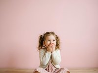 Young girl with curly blonde hair and fair complexion, wearing cream-colored long-sleeve top and pink skirt, sitting against soft pink background with hand near mouth in thoughtful or worried expression.